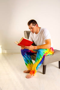 Thumbnail for Image of male model sitting on a chair reading a book wearing Rainbow Tie-Dye pajama lounge pants 