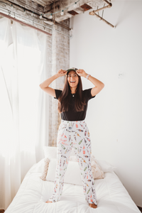 Thumbnail for Female model standing on a white bed facing forward smiling wide and wearing Just Screw It Wine pajama lounge pants