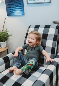 Thumbnail for Image of little girl model sitting on a plaid couch giggling wearing Sesame Street Oscar The Grouch pajama lounge pants 