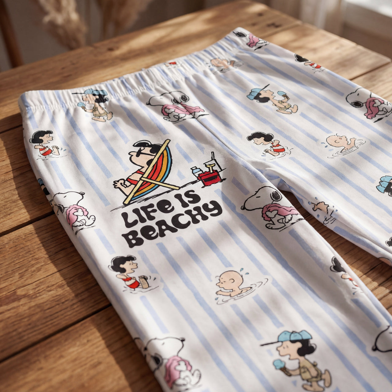 Close-up of Snoopy beach-inspired lounge pants on wooden surface, showcasing vibrant 'Life Is Beachy' graphic and breezy striped fabric.